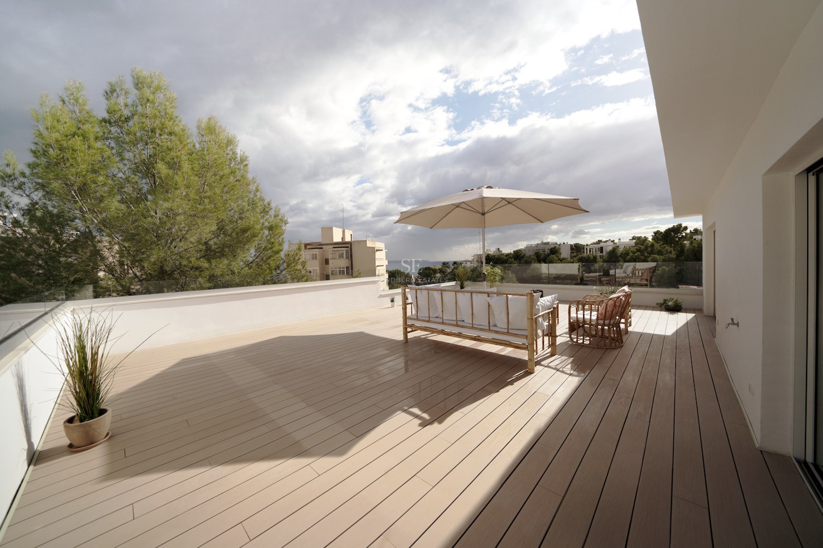 Terraza espaciosa con suelo de tarima, muebles de bambú, sombrilla beige y barandilla de cristal con vistas al mar.
