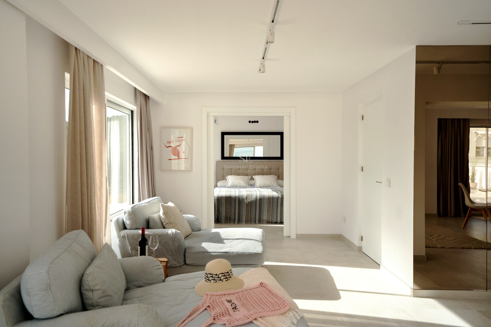 Suite de dormitorio moderna y luminosa con sofás azul claro, cortinas de lino y cama visible al fondo.
