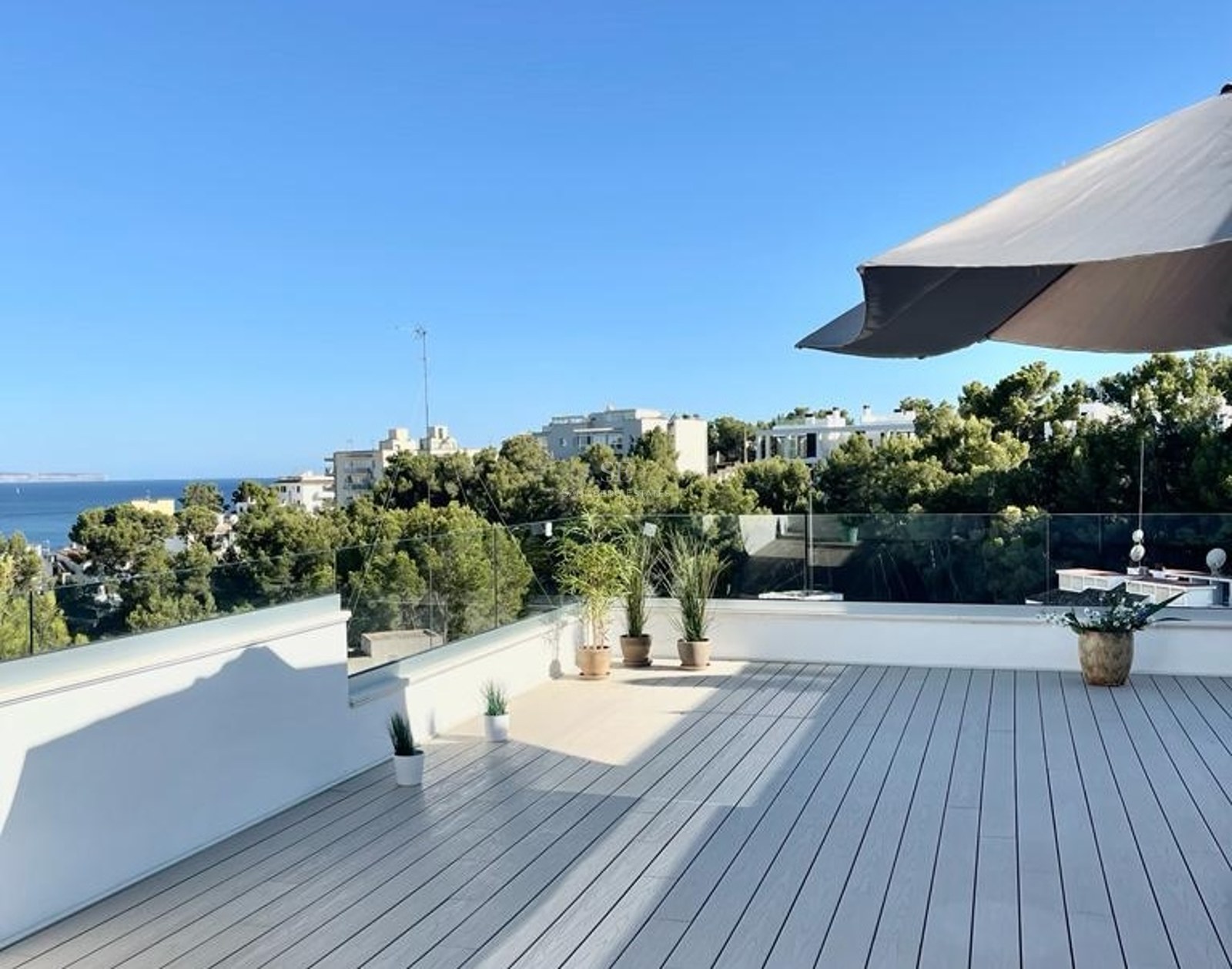 Terraza moderna con suelo de madera gris, barandillas de cristal y vistas panorámicas al mar y vegetación mediterránea.