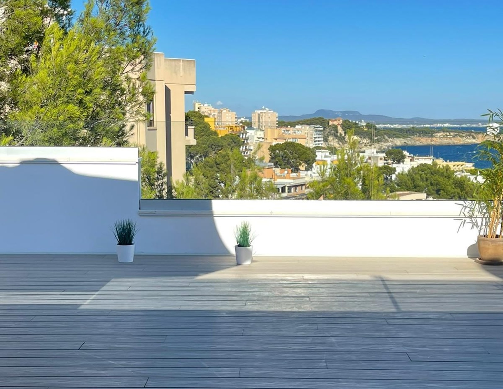 Terraza con suelo de madera gris y barandillas de cristal con vistas a la costa mediterránea y cielo azul despejado.
