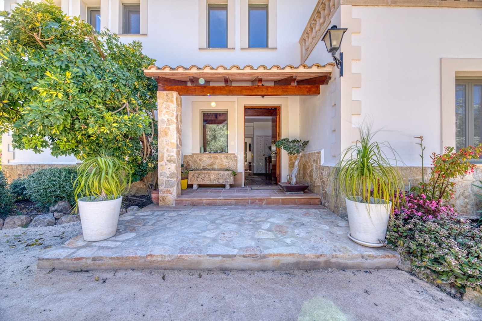Entrée de villa avec piliers en pierre, porche en bois, murs blancs et plantes en pots sous une lumière naturelle.