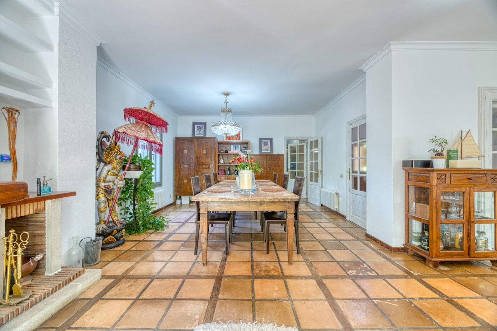 Grande salle à manger avec table en bois, sol en terre cuite, cheminée et statue décorative.