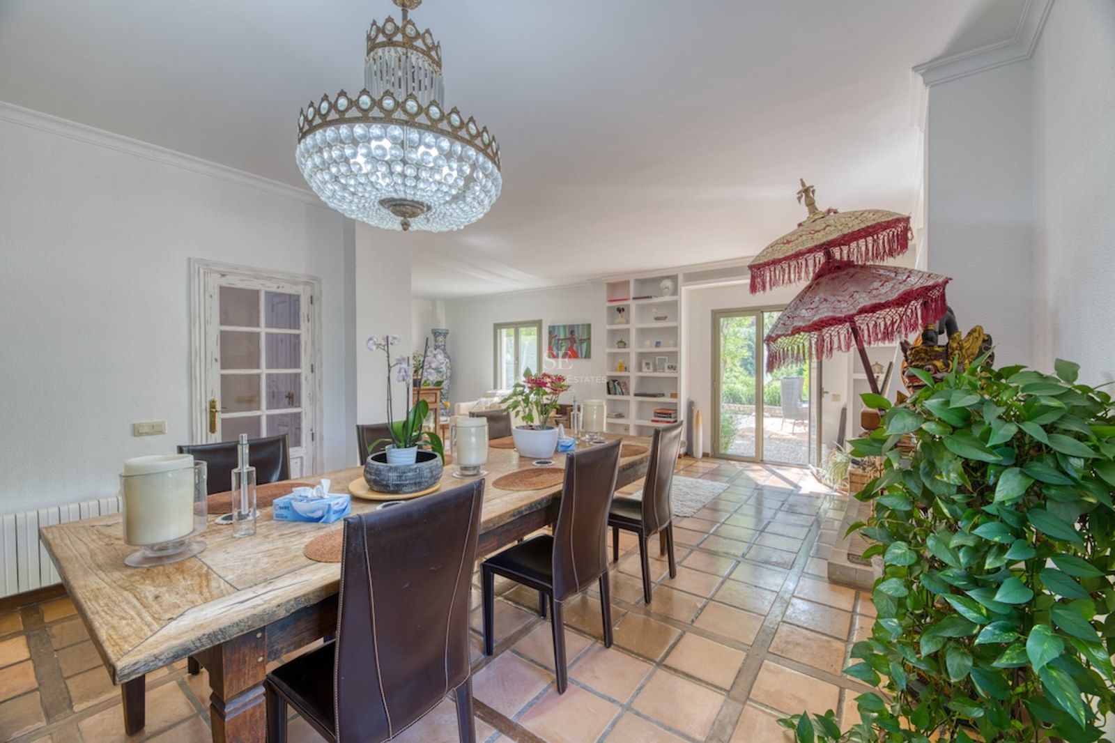 Salle à manger avec sol en terre cuite, table en bois rustique, chaises en cuir et lustre en cristal.