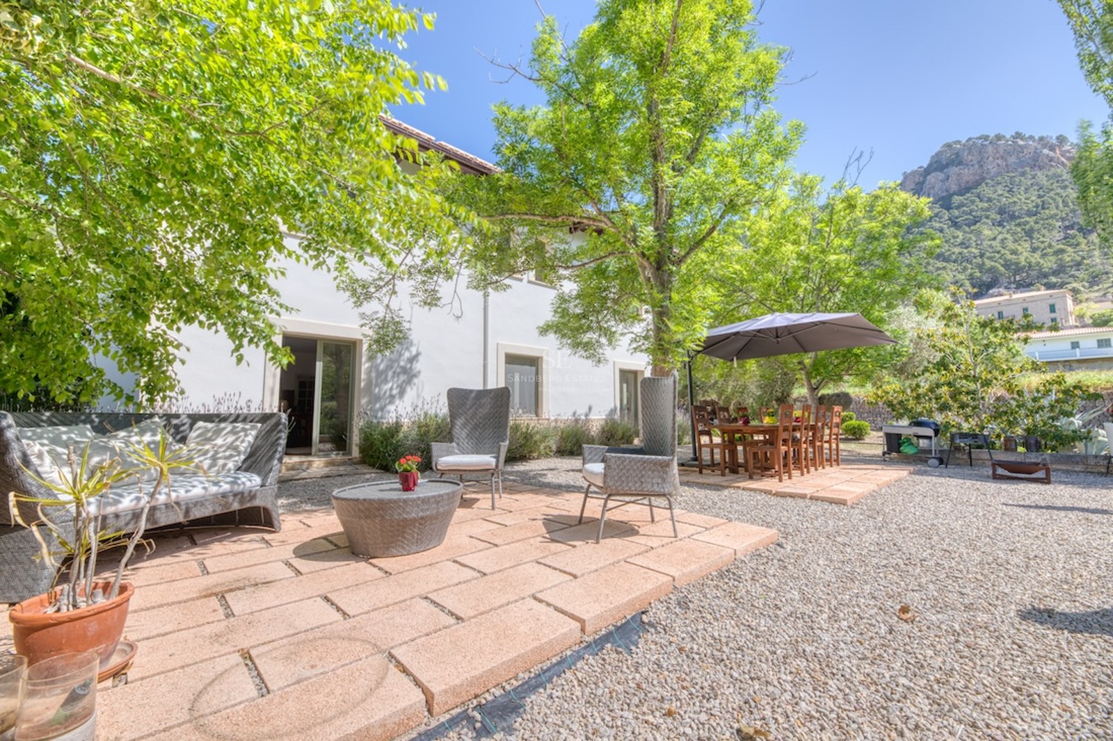 Terrasse ensoleillée avec salon en résine tressée et table à manger sous les arbres, face aux montagnes.