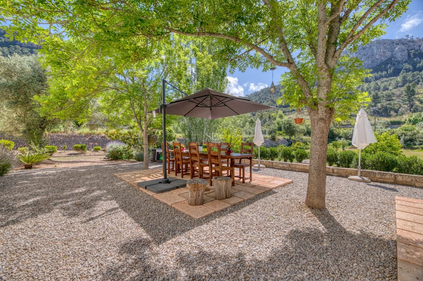 Table et chaises en bois sous un parasol sur une terrasse en gravier entourée d'arbres et de montagnes.