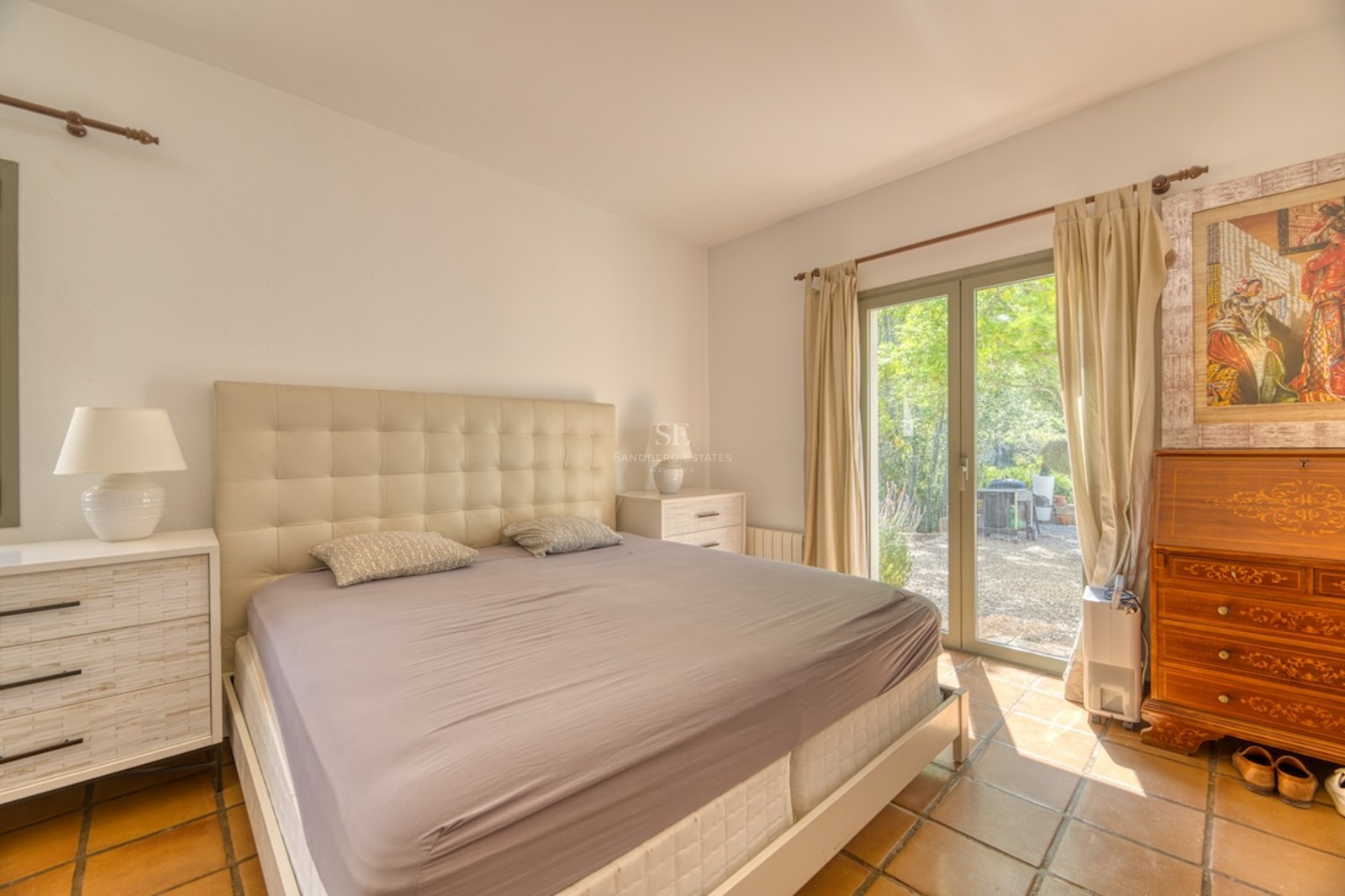 Chambre avec lit double, tête de lit capitonnée, sol en terre cuite et porte vitrée donnant sur le jardin.