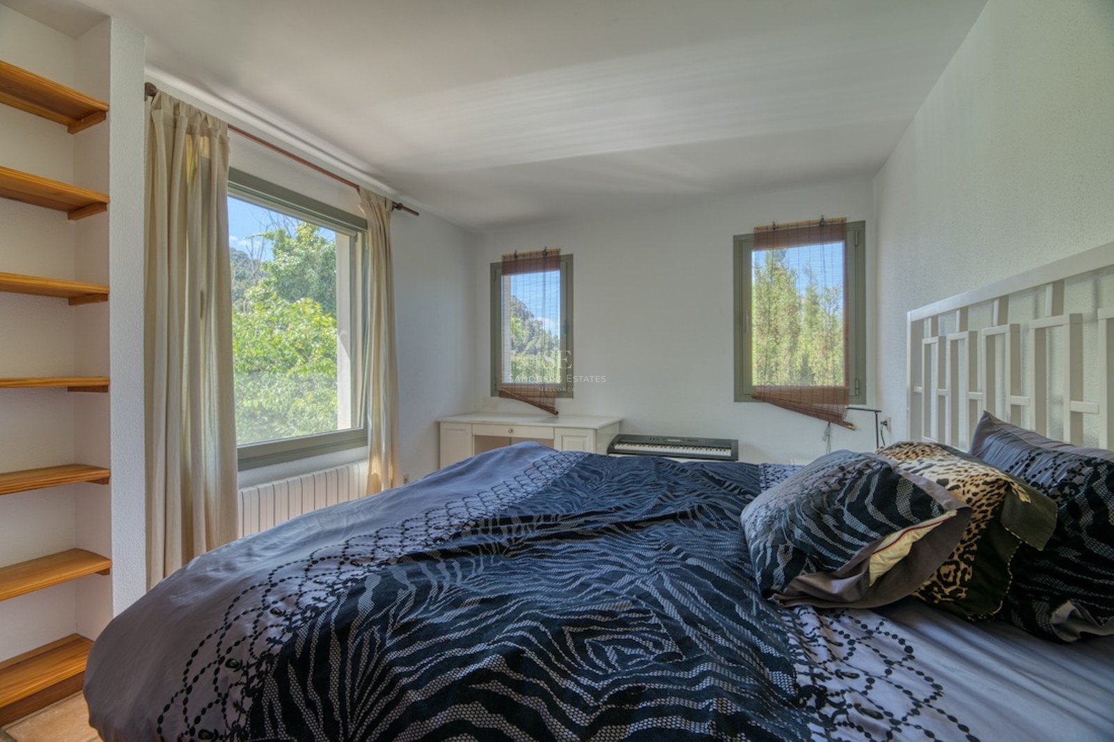 Chambre aux murs blancs, trois fenêtres donnant sur des arbres, lit avec coussins à motifs et étagères en bois.