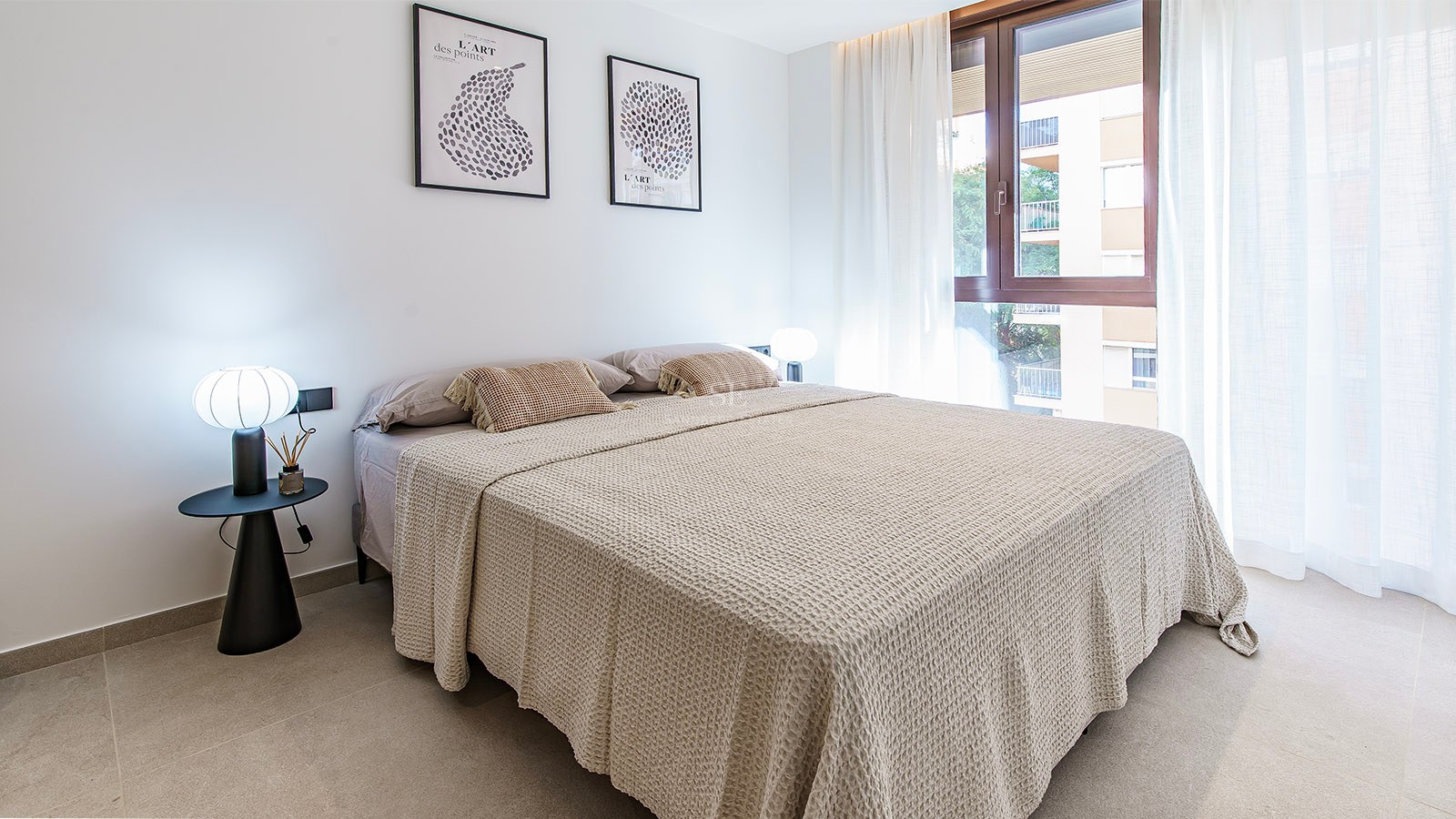 Ljust sovrum med stor säng, beige sängkläder, designerlampor och fönster med skira gardiner.