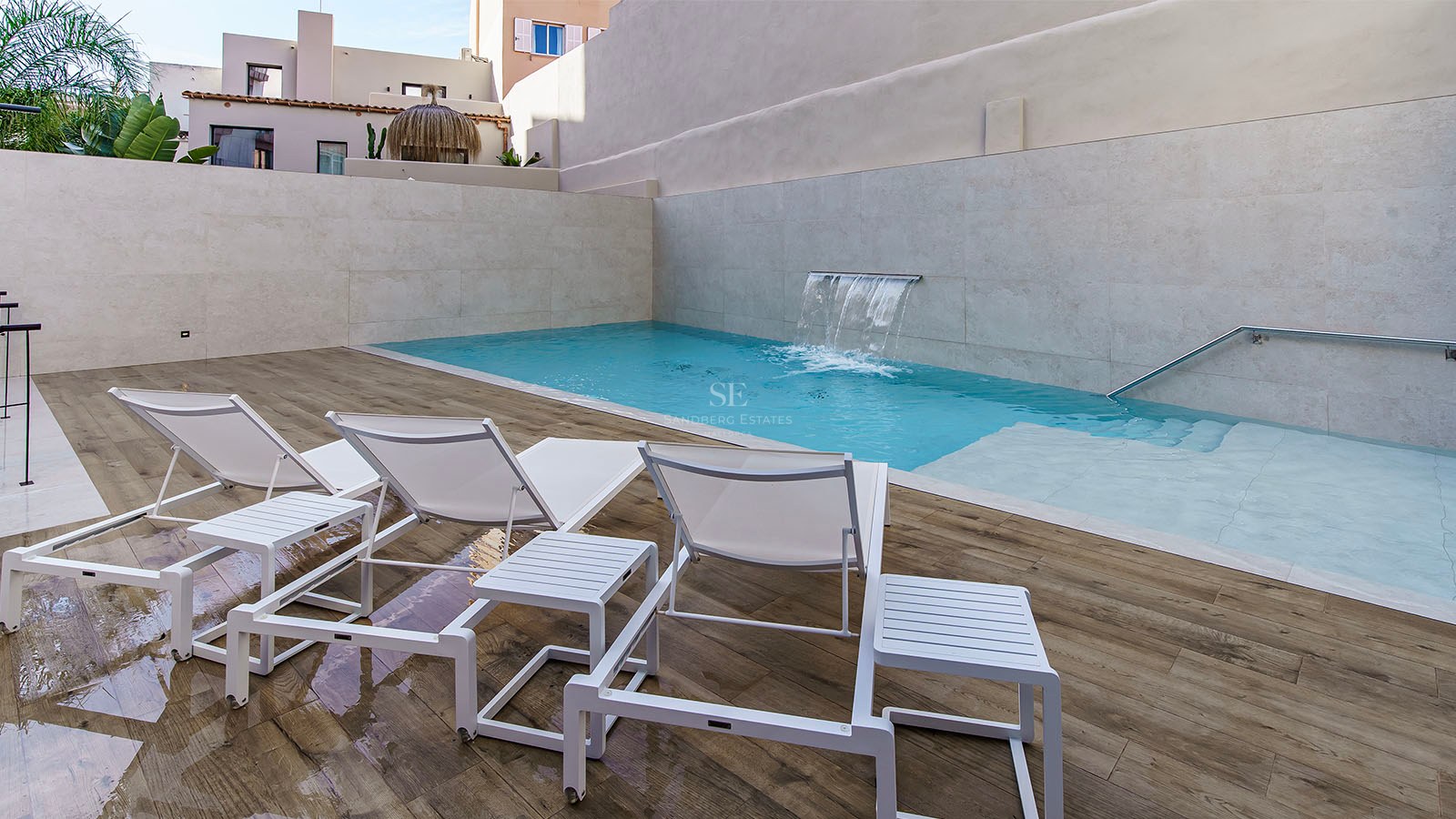 Turkos pool med integrerat vattenfall, vita solstolar på trädäck och stenväggar.
