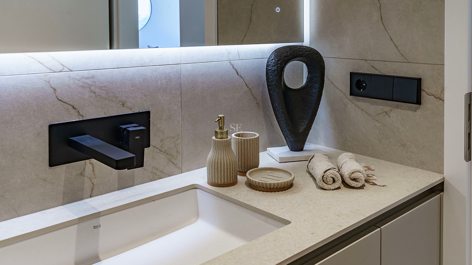 Närbild på ett vitt tvättställ med mattsvart kran, beige marmorvägg och designaccessoarer.