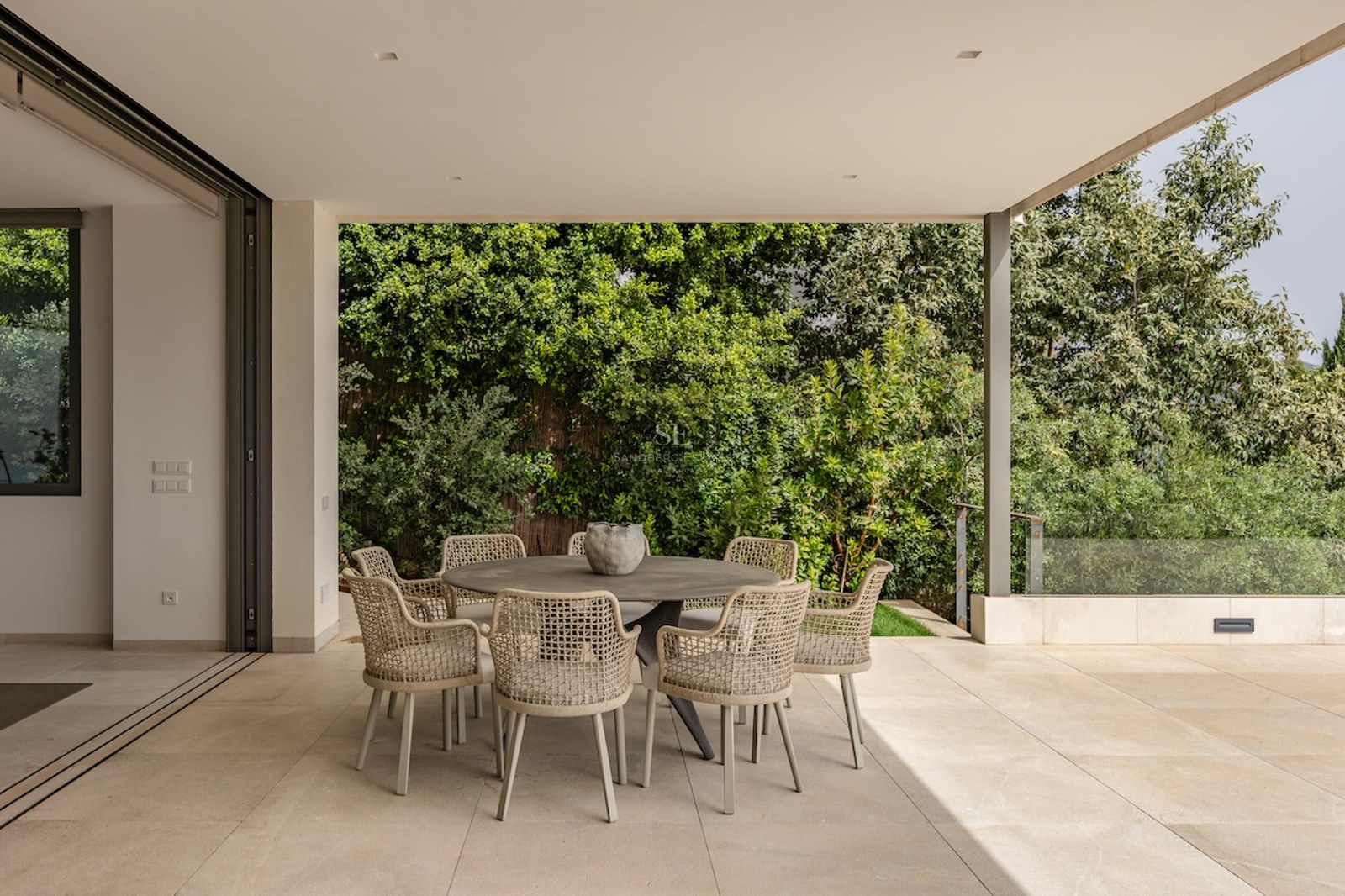 Modern covered terrace featuring a round dining table with woven chairs against a lush green garden backdrop.