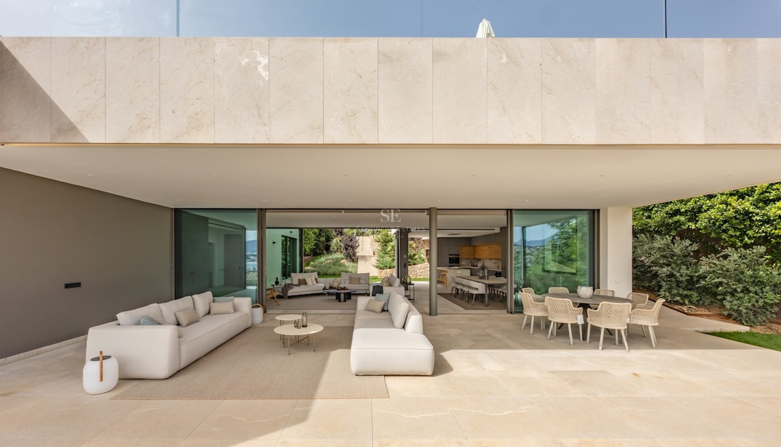 Luxury villa terrace featuring beige stone flooring, white sofas, and glass doors opening to living areas.