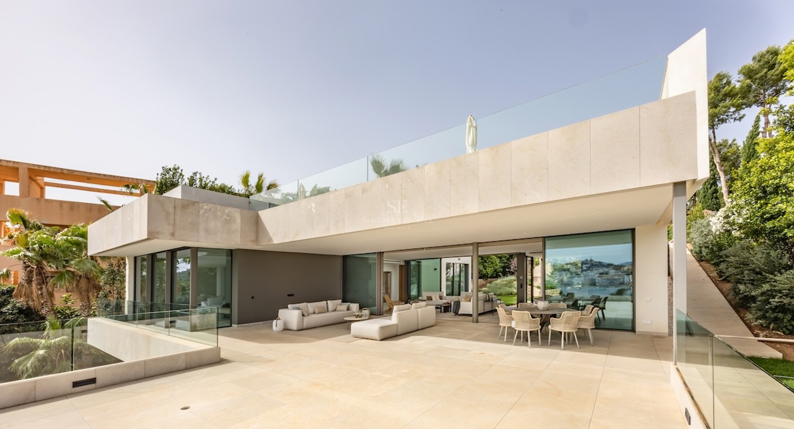 A wide sun-drenched stone terrace with modern lounge seating and a dining area under a large architectural overhang.