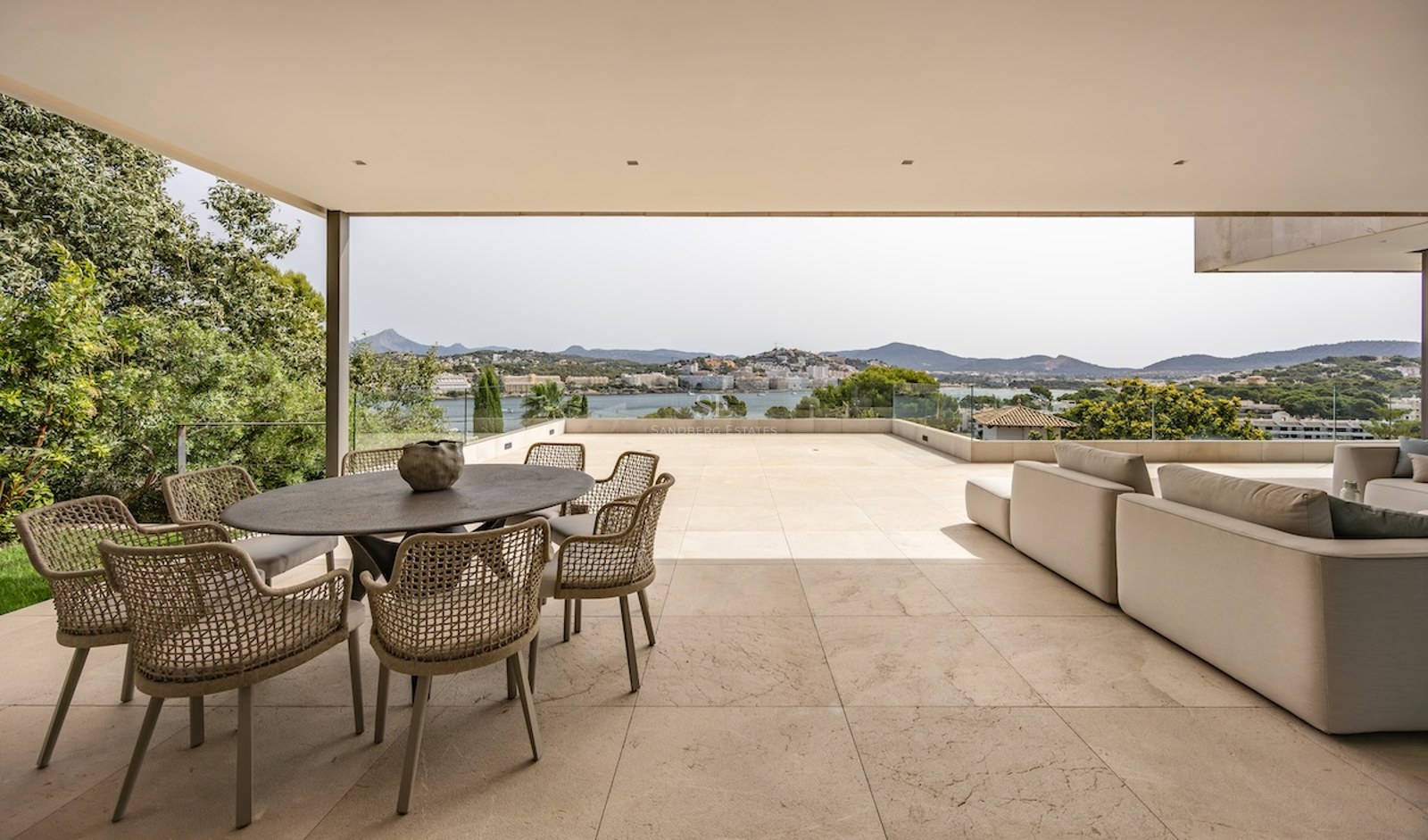 Covered terrace with stone flooring, outdoor dining set, and lounge sofa overlooking a bay and mountains.