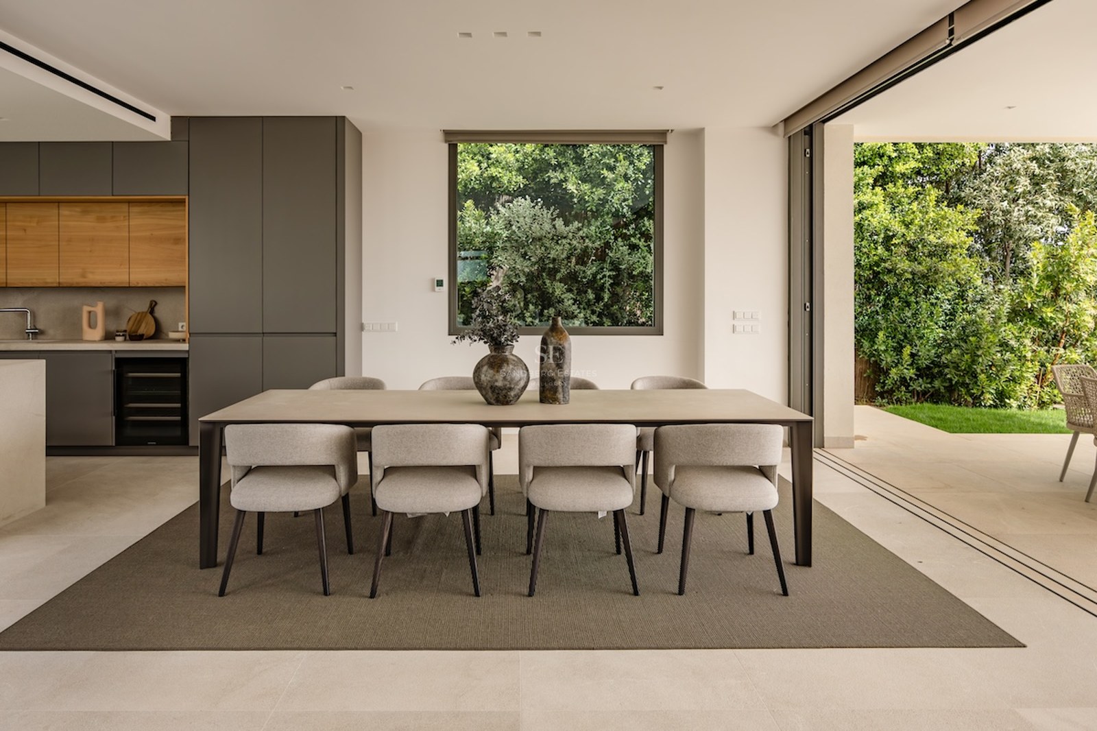 Modern dining room featuring a large rectangular table, eight beige upholstered chairs, and floor-to-ceiling glass doors.