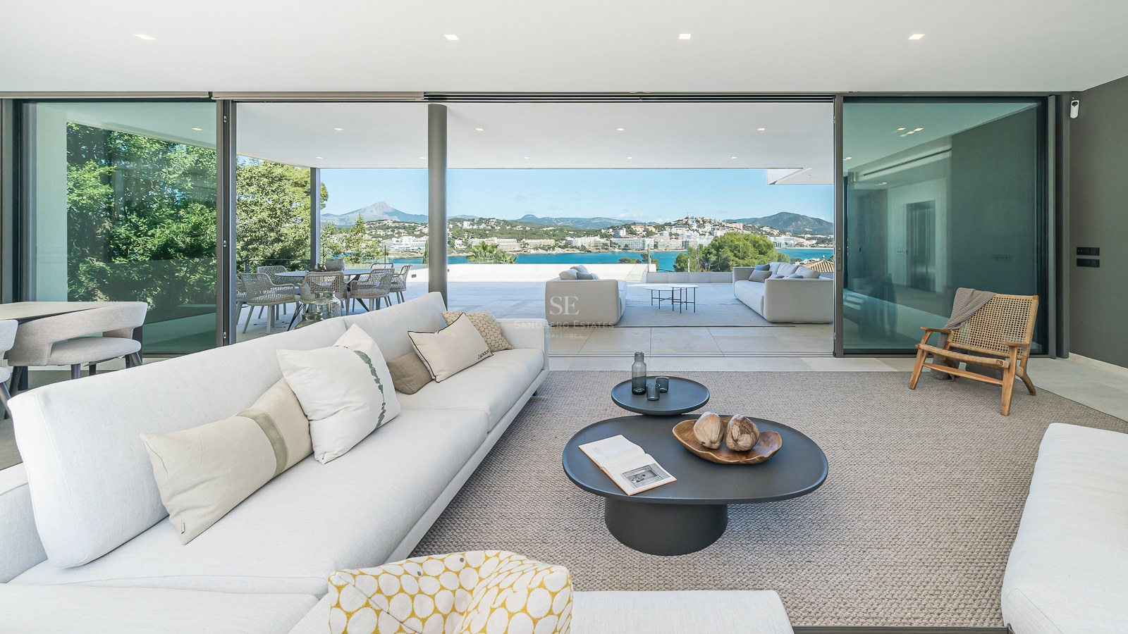 Modernt vitt vardagsrum med glasdörrar och terrass med panoramautsikt över havet och bergen.