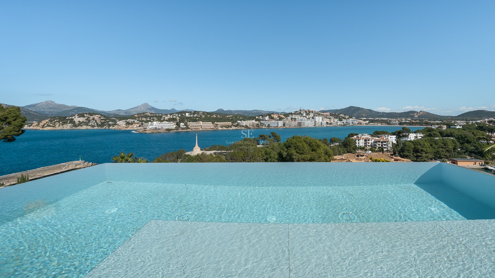 En infinitypool med utsikt över en blå vik, kustbyggnader och berg under en klar himmel.