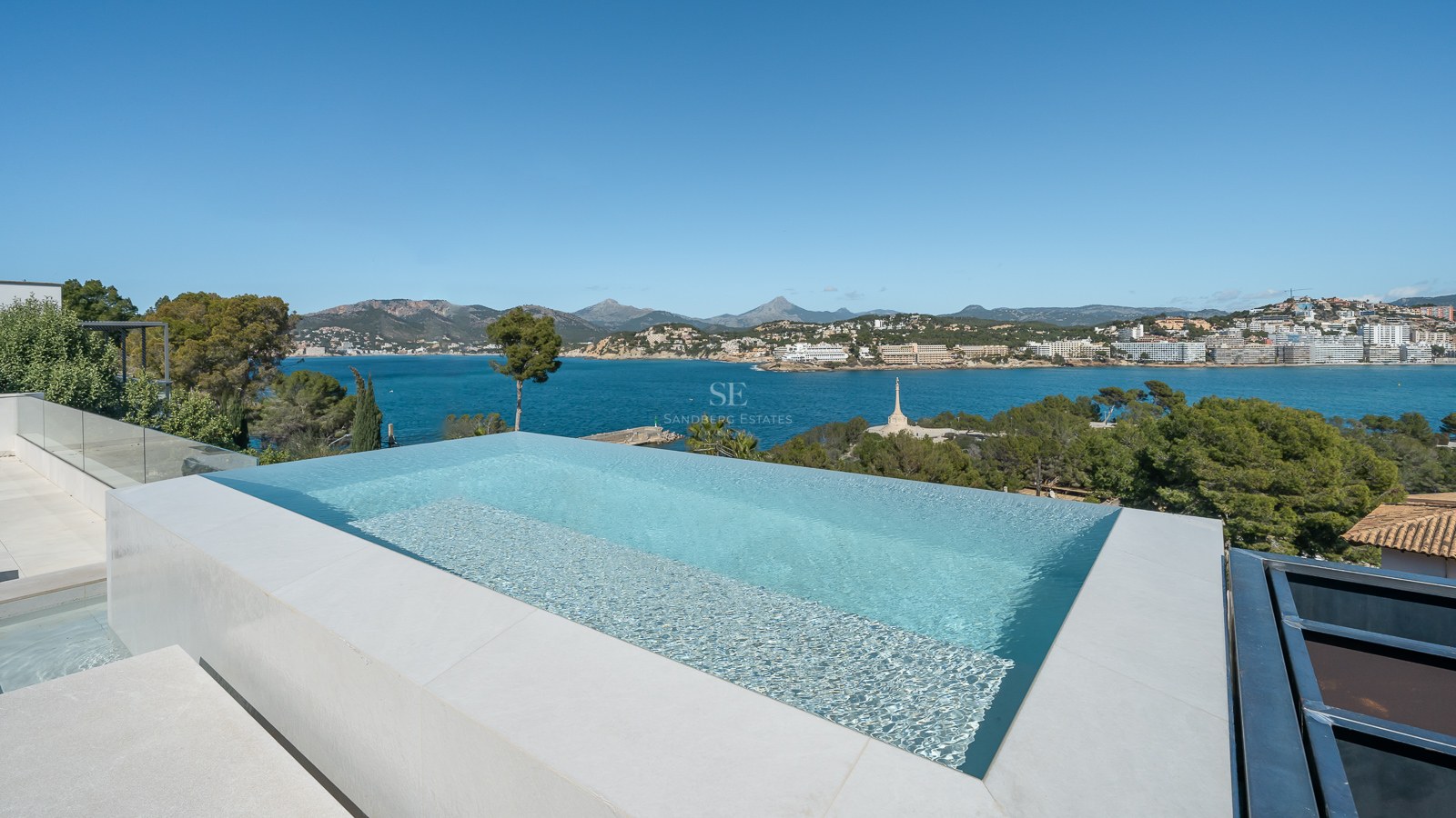 Modern infinitypool med turkost vatten som blickar ut över en blå vik, berg och kustlinje.