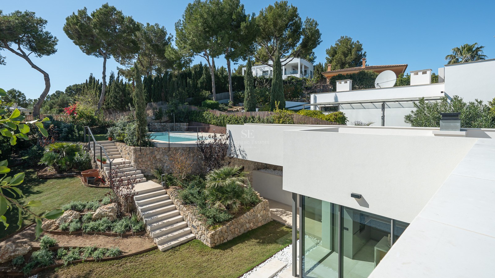 Modern vit villa med en terrasserad trädgård, stentrappor och en pool omgiven av tallar under en klarblå himmel.