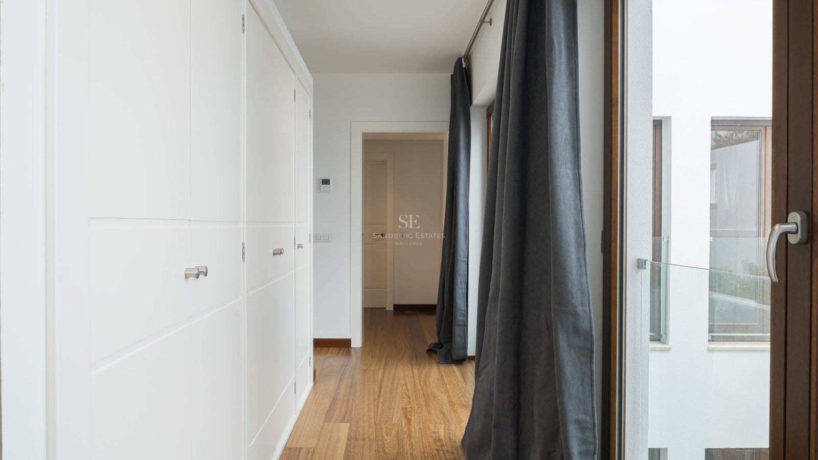 Pasillo luminoso con armarios empotrados blancos, suelos de madera cálida y cortinas de color gris marengo.