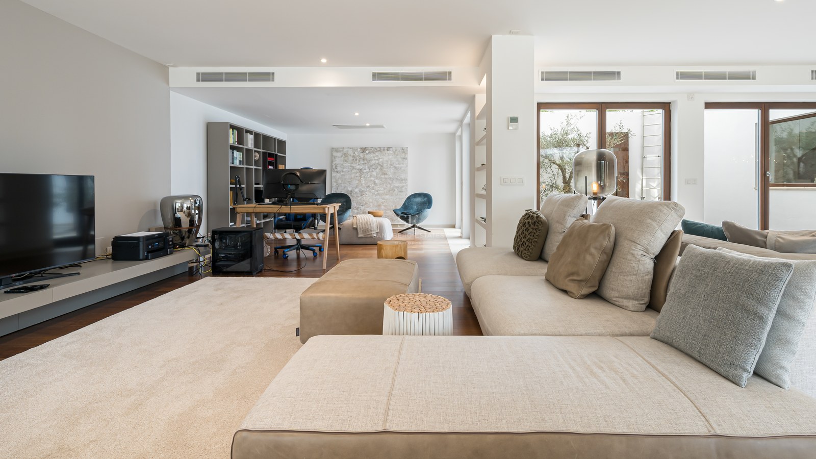 Salón moderno con sofá beige en forma de L, suelos de madera noble y zona de oficina al fondo con escritorio.