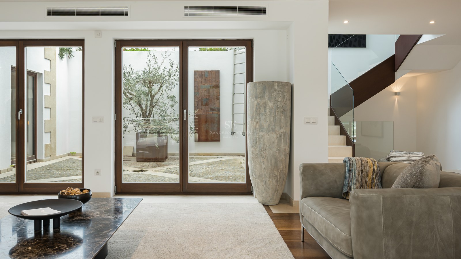 Salón moderno con sofá de cuero, mesa de mármol y grandes puertas de cristal que dan a un patio con un olivo.