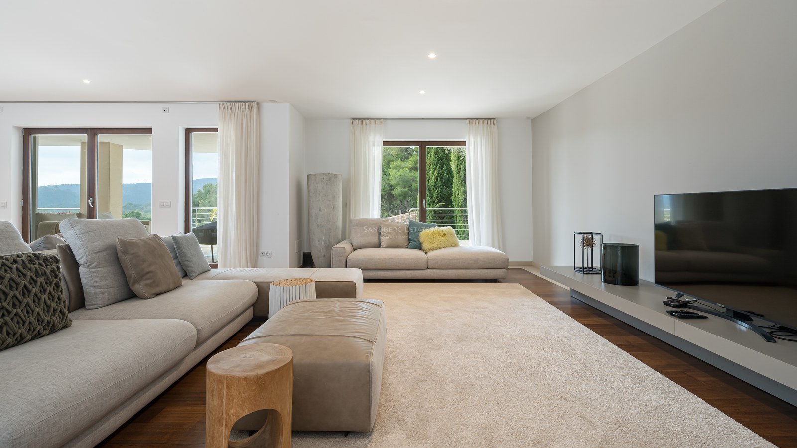 Amplio salón con sofás beige, otomana de cuero, suelos de madera oscura y ventanales con vistas a colinas verdes.