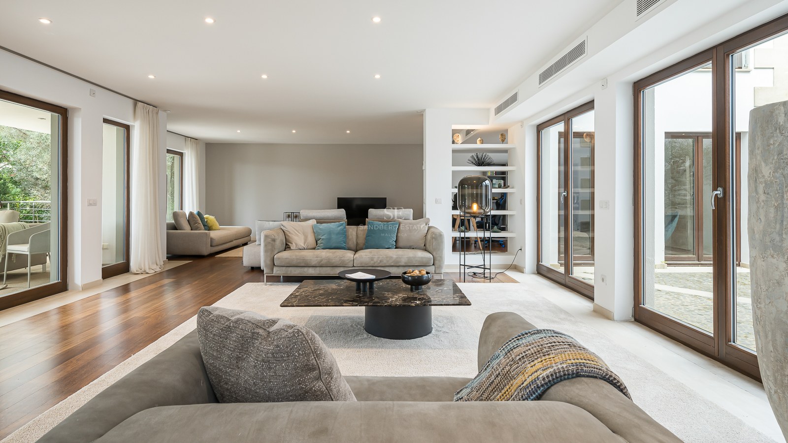Amplio salón moderno con suelos de madera, sofás grises, mesa de mármol y grandes ventanales de cristal.