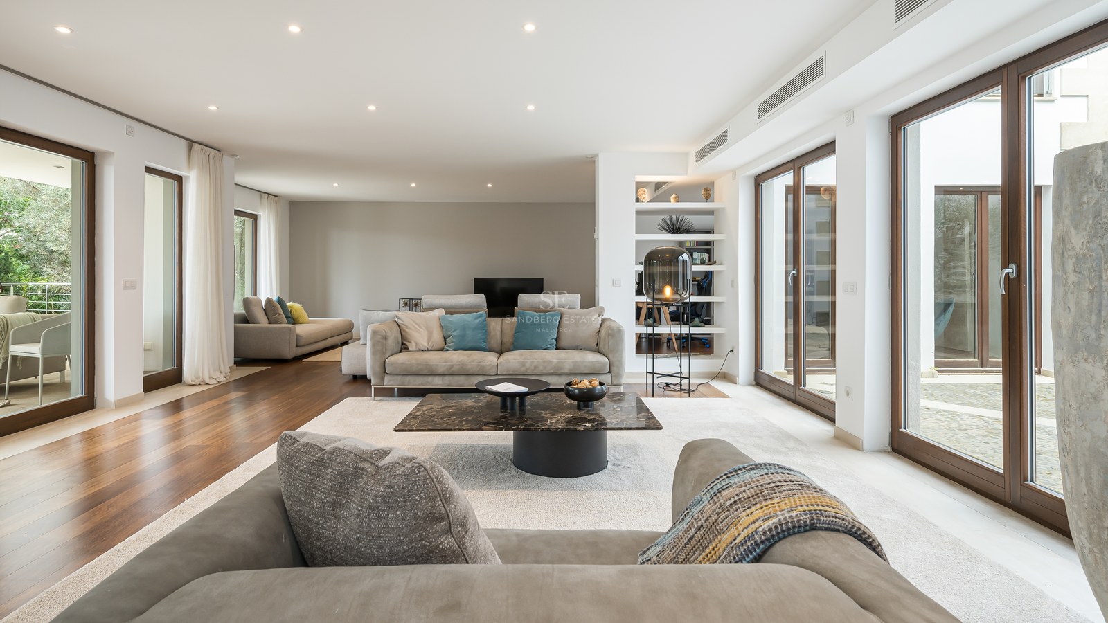 Amplio salón moderno con suelos de madera, sofás grises, mesa de centro de mármol y ventanales de suelo a techo.