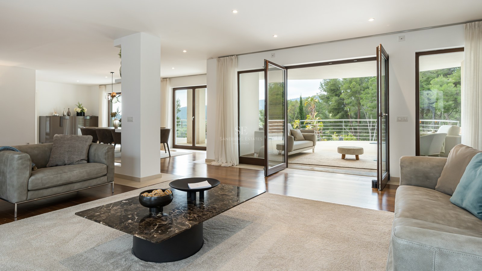 Salón moderno con sofás de cuero, mesa de mármol y grandes puertas de cristal que dan a una terraza con vistas al bosque.