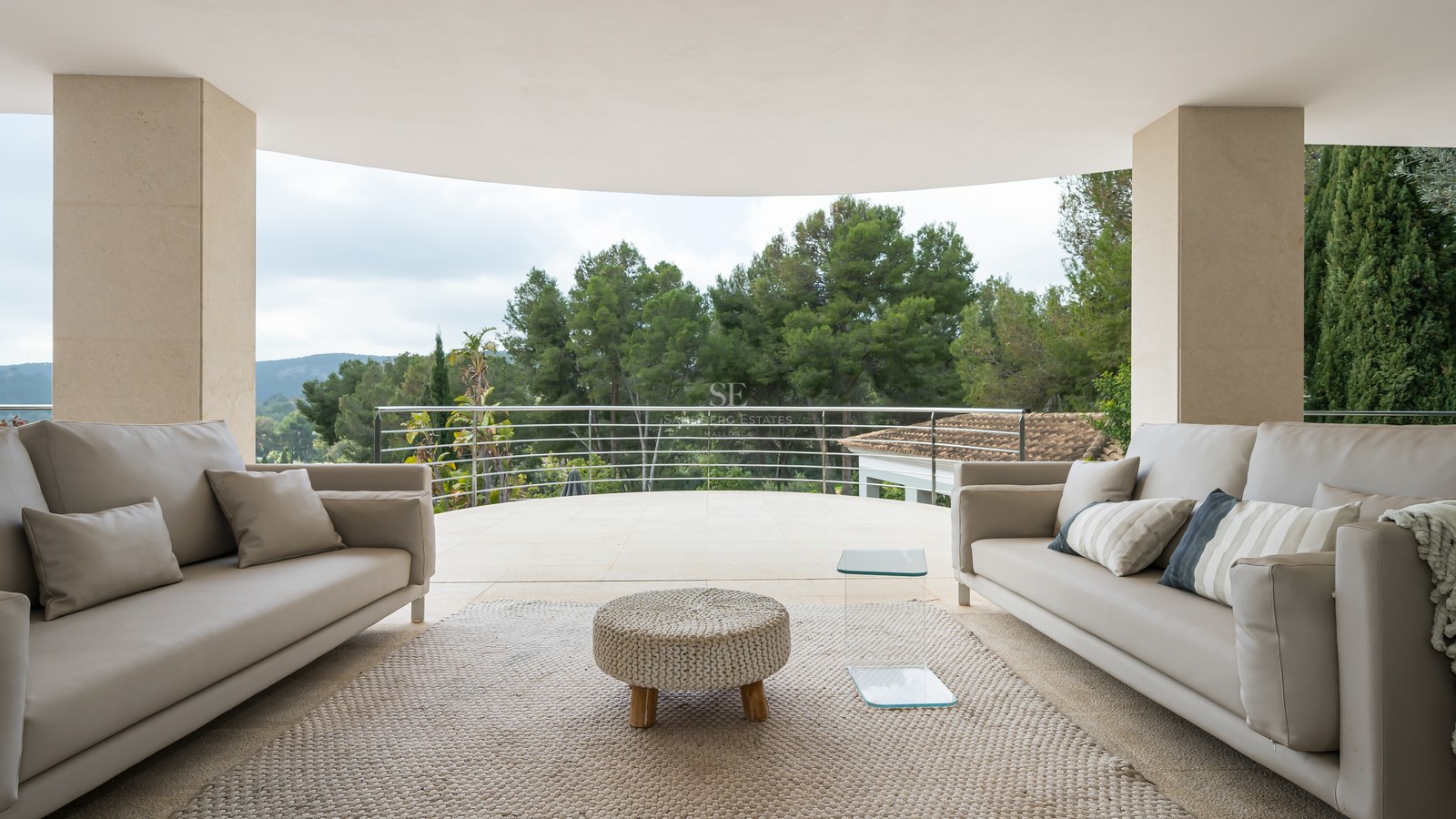 Terraza moderna con sofás grises y puf tejido frente a un bosque denso bajo un cielo despejado.