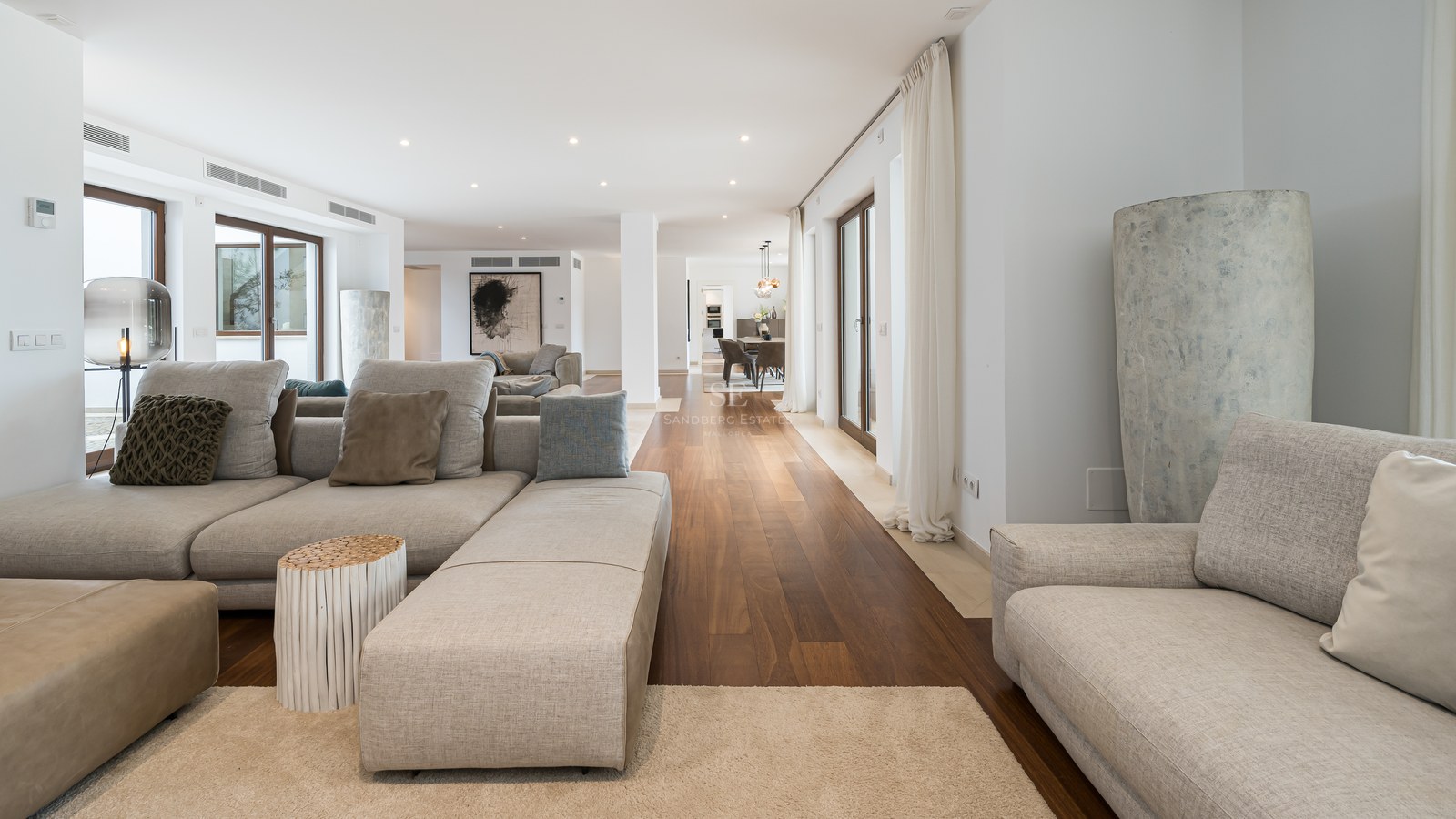 Amplio salón moderno con sofás de tela gris, suelos de madera oscura y grandes ventanales de cristal.