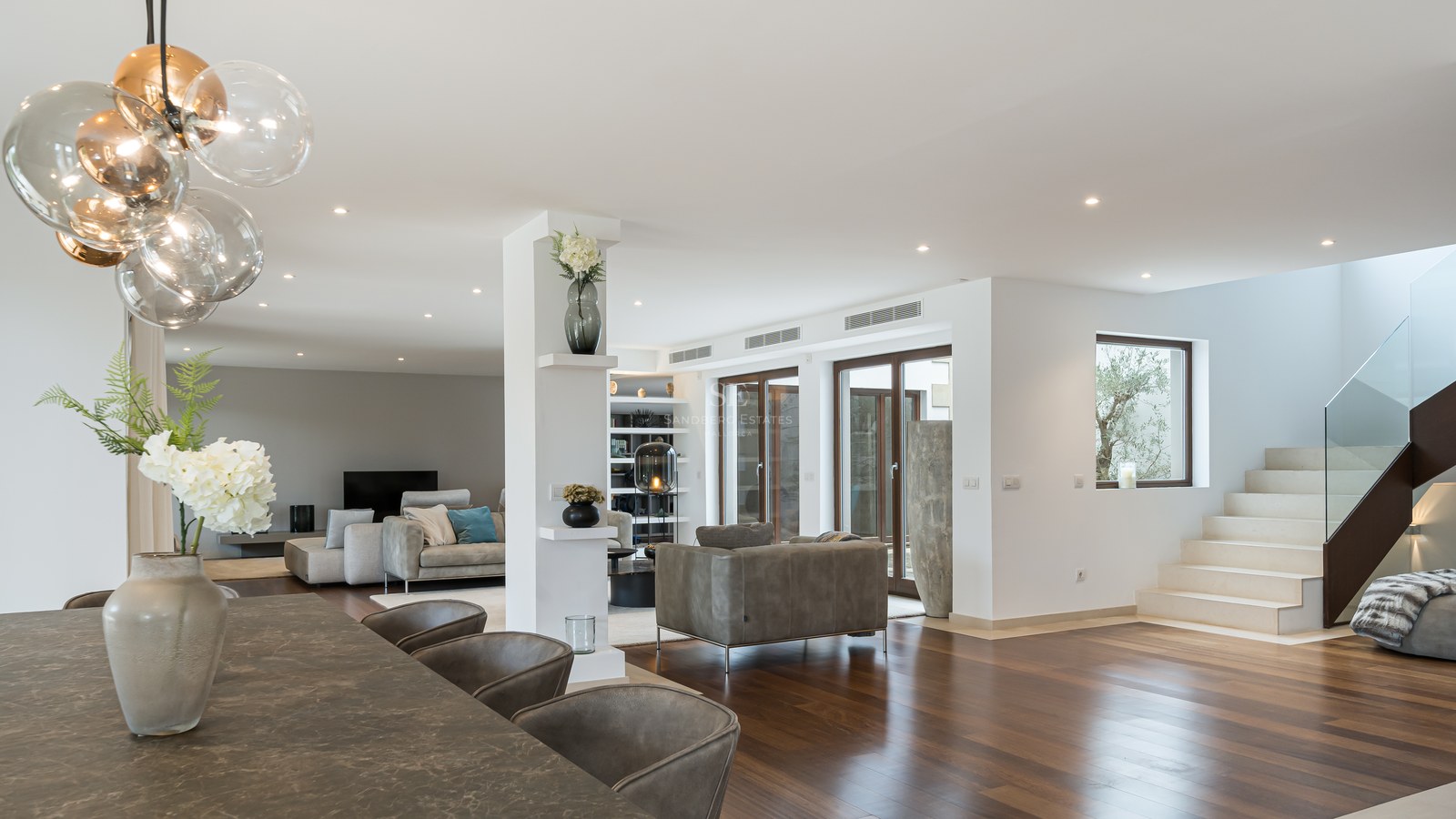 Amplio salón con suelos de madera oscura, mesa de piedra, lámpara de esferas de cristal y escalera de piedra natural.