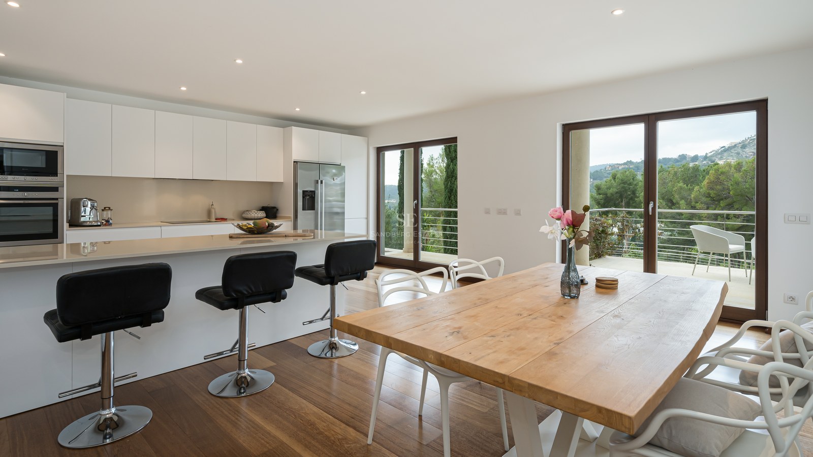 Cocina blanca contemporánea con barra americana, mesa de comedor de madera y ventanales con vistas al exterior.