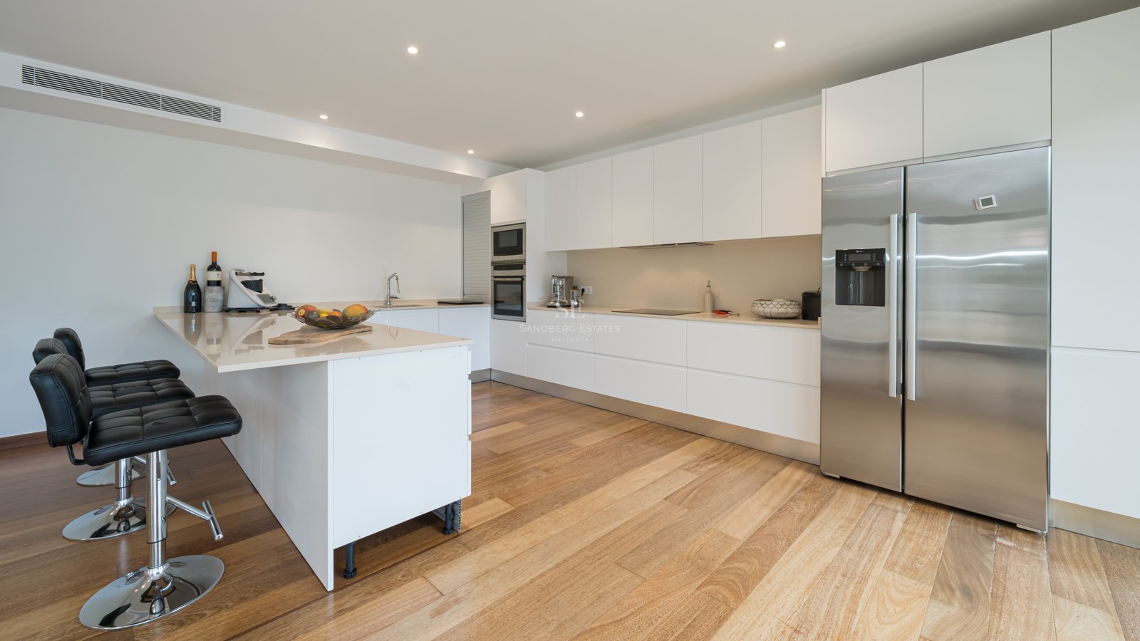 Cocina blanca con suelo de madera, frigorífico de acero inoxidable, encimera clara y taburetes de cuero negros.