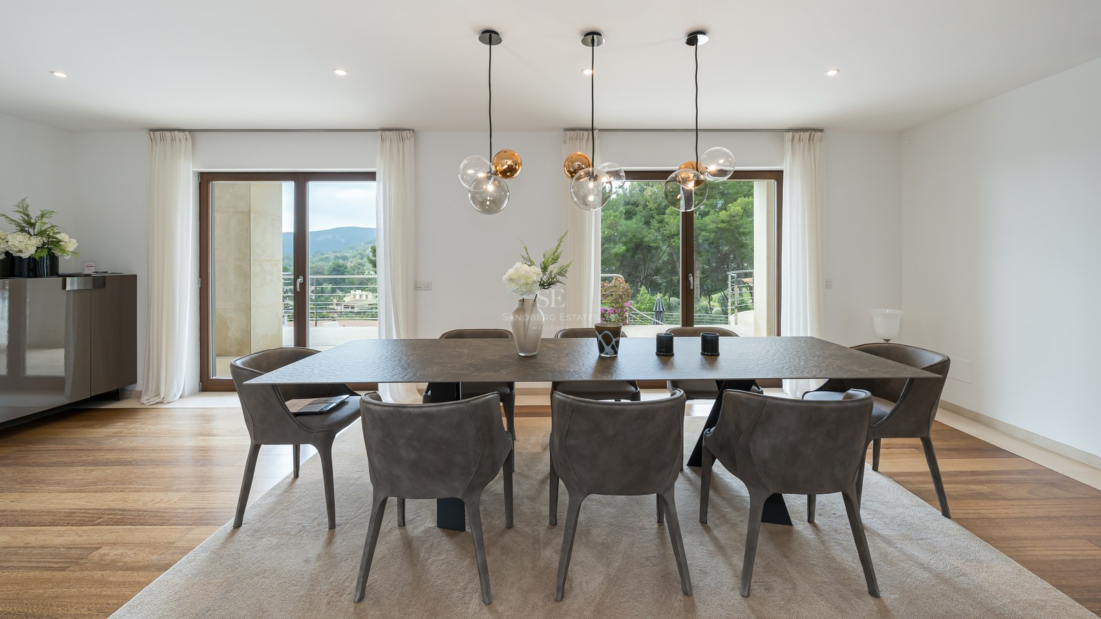 Zona de comedor moderna con mesa oscura texturizada, sillas grises, lámparas de diseño y grandes ventanales al exterior.