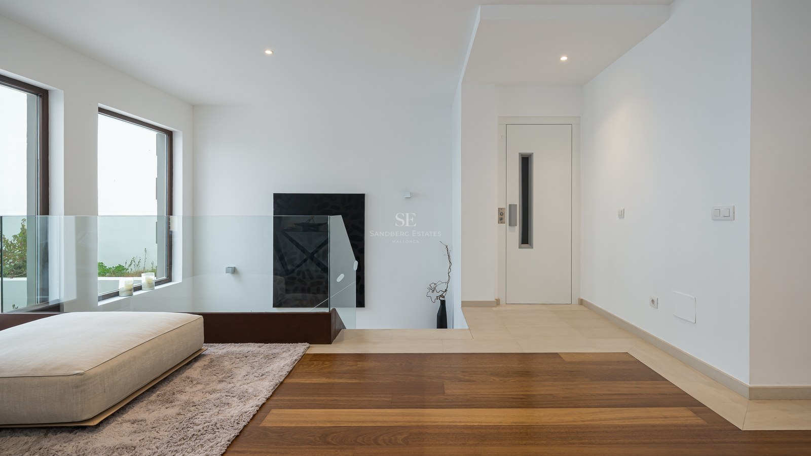 Distribuidor moderno con barandilla de cristal, suelo de madera de roble y puerta de ascensor blanca.