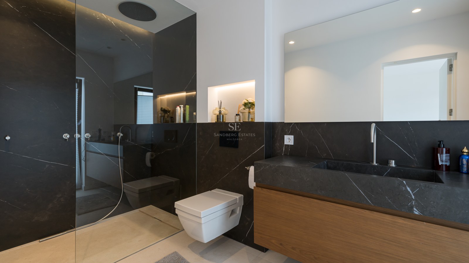 Baño moderno con paredes de mármol negro, inodoro suspendido, mueble de madera y ducha de obra acristalada.