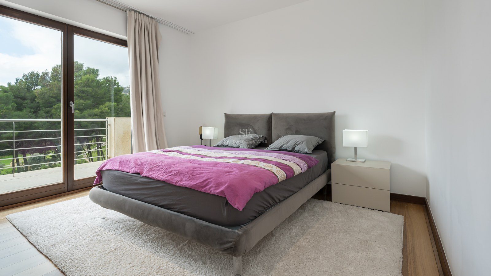 Dormitorio contemporáneo con cama tapizada gris, edredón púrpura, alfombra blanca y puerta de cristal a un balcón.