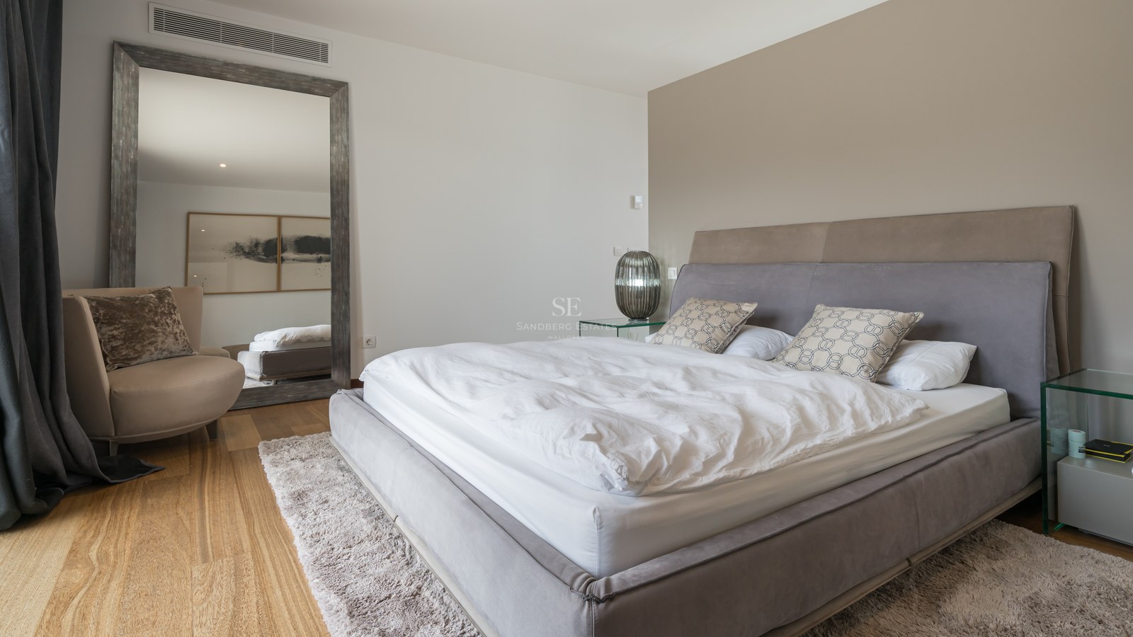 Dormitorio moderno con cama tapizada gris, suelo de roble, espejo de pie grande y mesitas de noche de cristal.