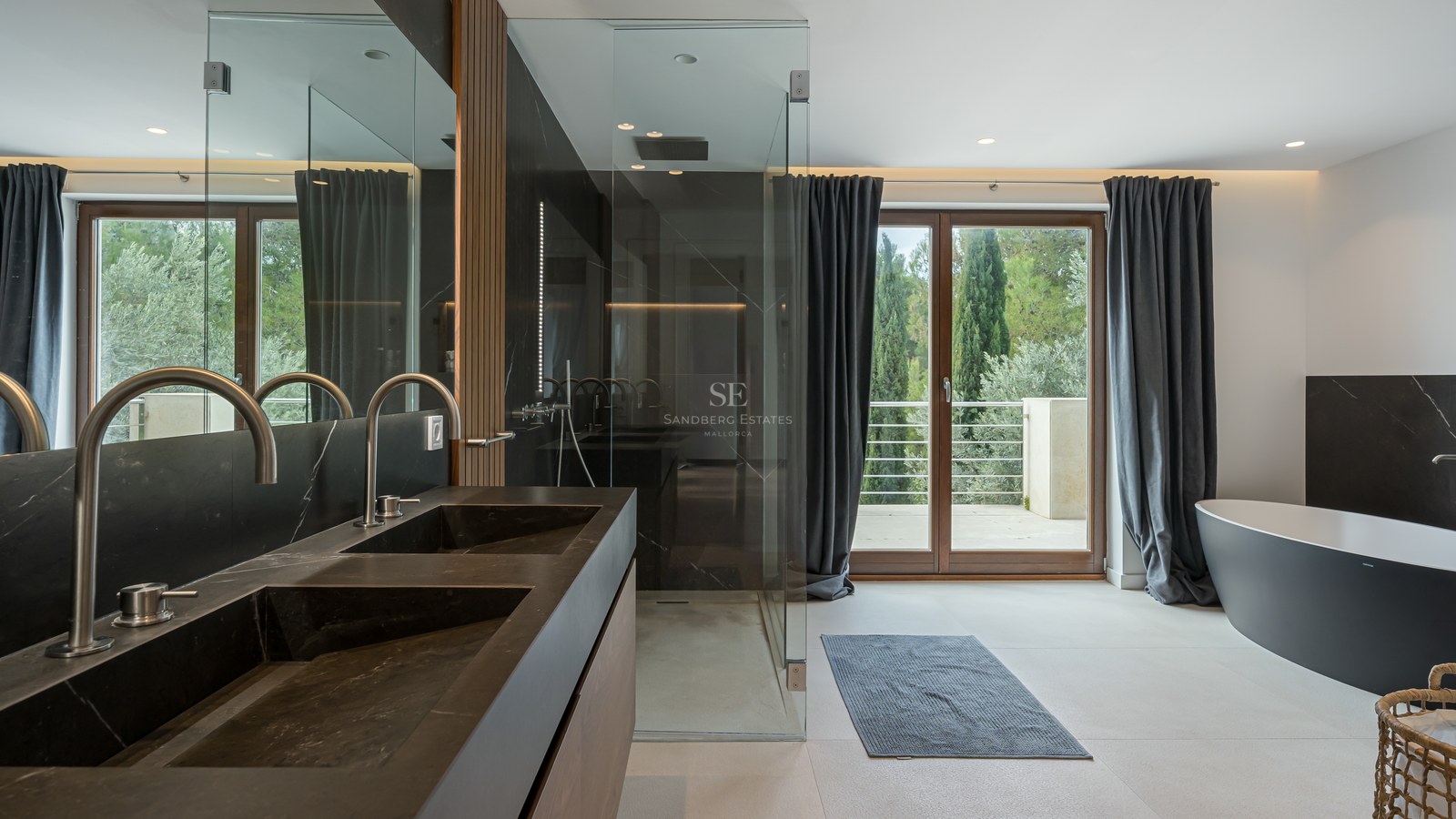 Baño moderno con lavabo doble negro, ducha de cristal, bañera exenta y acceso a terraza con vistas a la naturaleza.