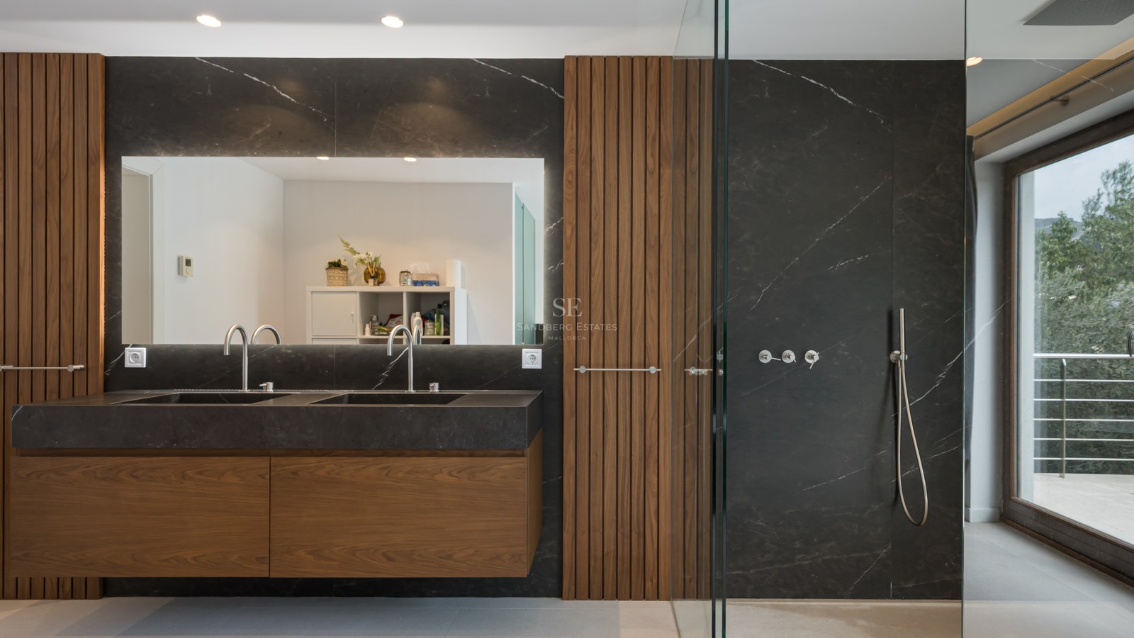 Baño principal moderno con lavabo doble de mármol negro, paneles de madera de nogal y amplia ducha de cristal.