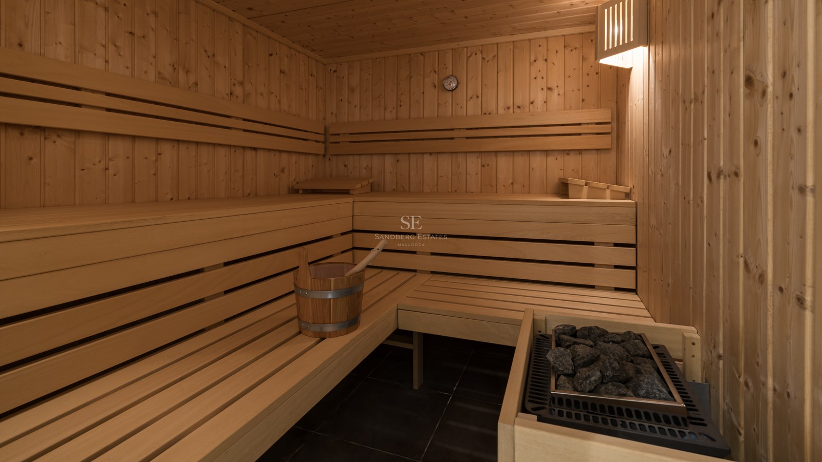 Vista interior de una sauna de madera con asientos a varios niveles, piedras térmicas y un cubo de madera.