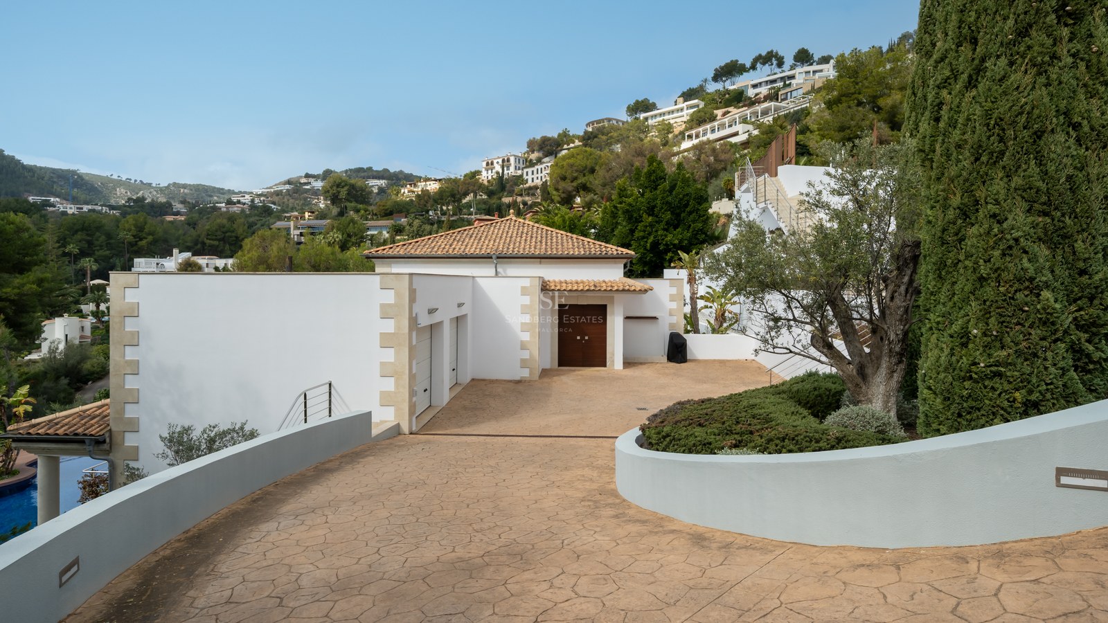 Entrada privada pavimentada hacia una villa blanca con tejado de terracota, garaje doble y vegetación mediterránea.