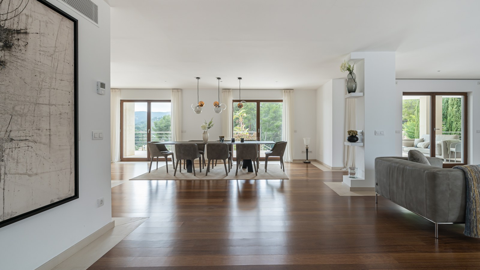 Comedor espacioso con suelos de madera oscura, lámparas de diseño y grandes ventanales hacia un jardín frondoso.