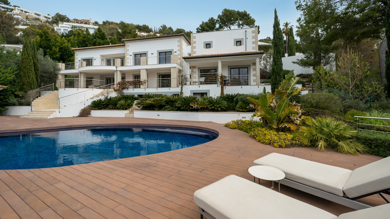 Villa de lujo blanca con piscina curva, amplia terraza de madera y tumbonas frente a una fachada moderna.