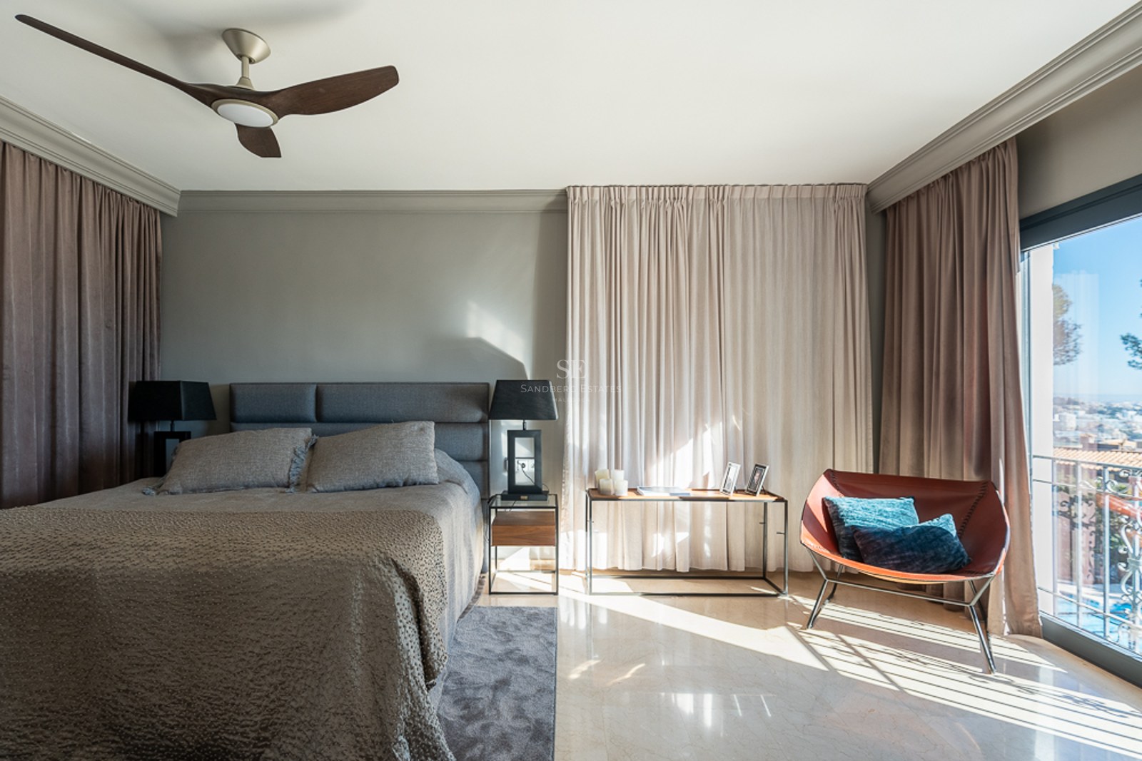Dormitorio moderno con cama tapizada gris, suelo de mármol, sillón de cuero y balcón con vistas a la costa.
