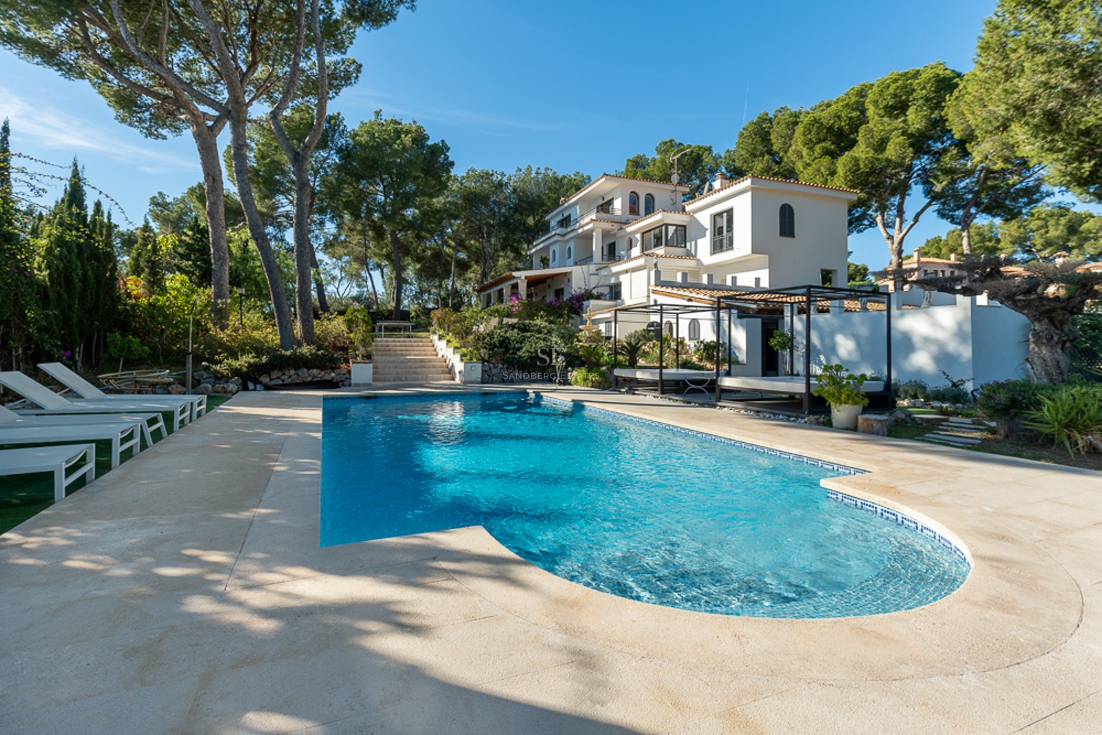Amplia piscina azul con terraza de piedra, tumbonas blancas y villa de varios niveles rodeada de pinos.