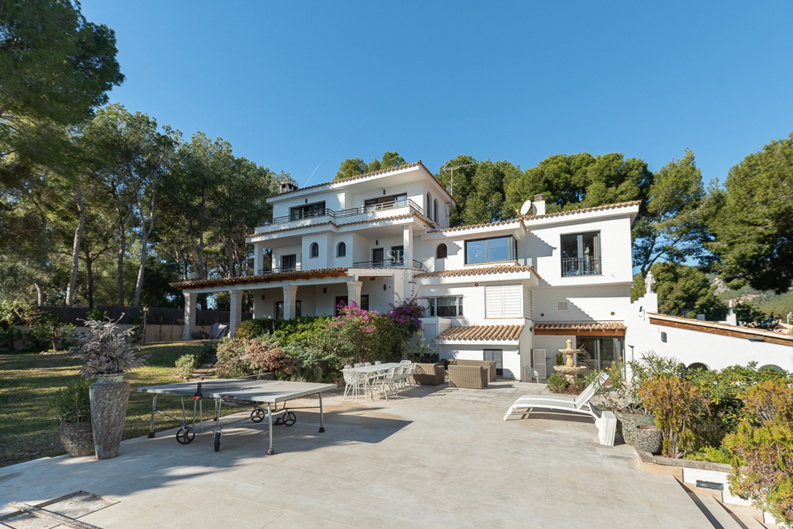 Vista exterior trasera de una villa mediterránea blanca de tres niveles con terrazas de piedra y entorno boscoso.