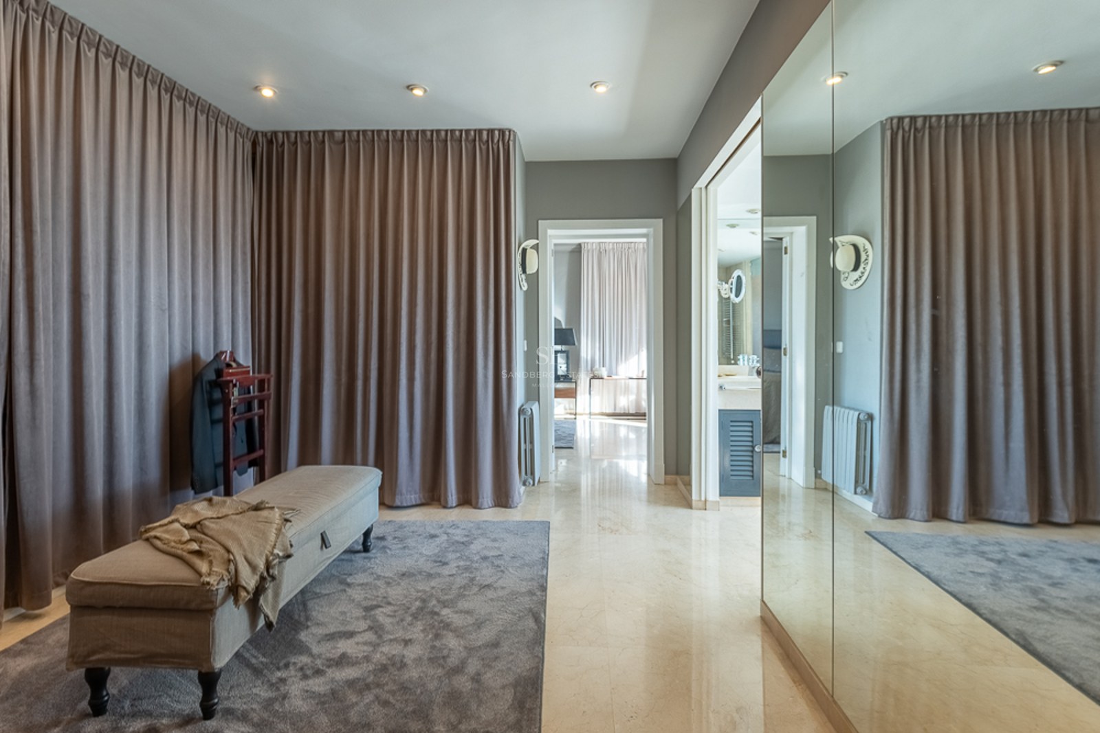 Vestidor moderno con suelos de mármol pulido, cortinas de suelo a techo, banco tapizado y grandes espejos.